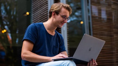 Mann sitzt vor der Universität und schaut in seinen Laptop.