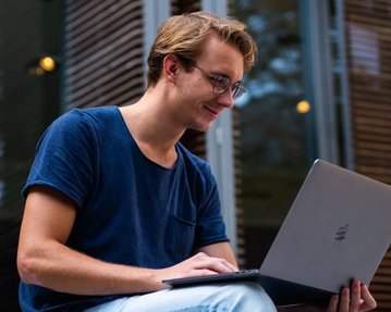 Mann sitzt vor der Universität und schaut in seinen Laptop.