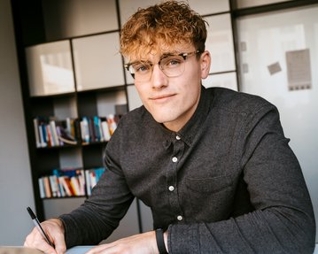 Young man with curly hair and glasses, sitting at a table and writing with a pen. Bookshelves are visible in the background.