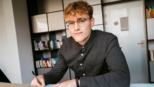 Young man with curly hair and glasses, sitting at a table and writing with a pen. Bookshelves are visible in the background.