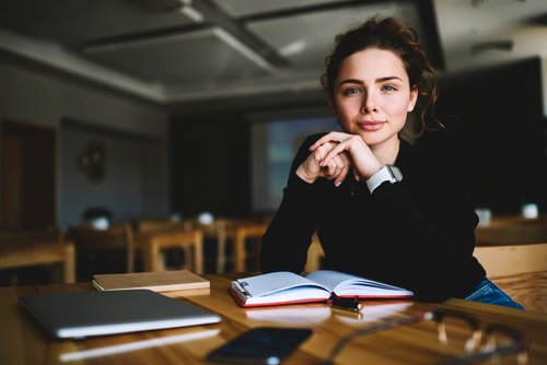 Junge Frau mit lockigem Haar sitzt an einem Tisch, schaut in die Kamera und hat ein Notizbuch sowie einen Laptop vor sich.