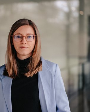 A young woman with long, straight hair and glasses stands in front of a modern glass wall. She is wearing a light blue blazer and a black top, smiling warmly at the camera.