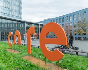Large, orange letters spell out the word "core" outdoors, surrounded by grass and a modern building in the background.