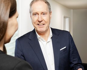 Ein Mann in einem Anzug spricht mit einer Person, die ihm zugewandt ist. Der Mann hat graue Haare und lächelt freundlich.
