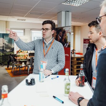 Three men are standing at a table, one is pointing at something with his finger. They all have name tags on and are engaged in discussion. Drinks are on the table.