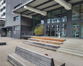 Eingangsbereich eines modernen Gebäudes mit Treppen, Holzbank und Pflanzen. Die Fassade ist aus grauem Material und hat große Fenster.
