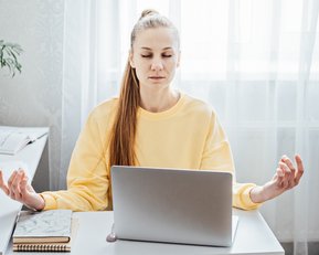 Eine Frau in einem gelben Sweatshirt sitzt an einem Schreibtisch und meditiert, während sie vor einem Laptop sitzt. Pflanzen sind im Hintergrund.