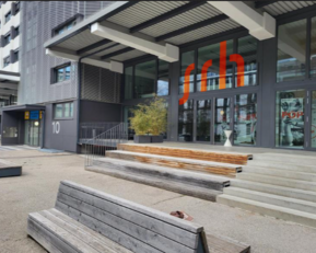 Entrance of a modern building with large windows and a striking red sign reading 'srh'. In front of the building, there are wooden steps and a bench.