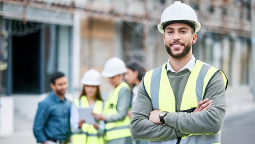 A construction manager in safety gear and a helmet stands smiling in the foreground, while in the background, other workers are engaged in a project.