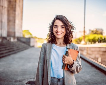 Eine junge Frau mit lockigem Haar trägt einen Blazer und hält ein Notizbuch sowie einen Ordner. Sie steht vor einem architektonischen Hintergrund und lächelt freundlich in die Kamera.