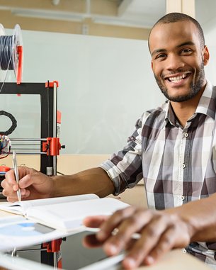 A man sits at a table with a 3D printer, a notebook, and a tablet, holding a pen and smiling.
