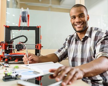 A man sits at a table with a 3D printer, a notebook, and a tablet, holding a pen and smiling.