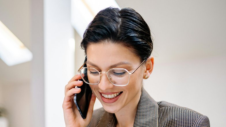 A smiling woman with glasses is talking on the phone while taking notes on a document. She is wearing a checked blazer and sitting at a table in a bright room.