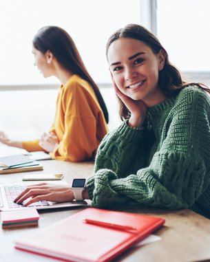 A young woman with long brown hair sits at a table, smiling as she works on a laptop. In the background, two other people are also working on laptops. Red notepads are spread out on the table.