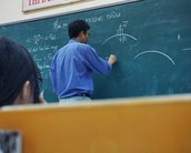 A teacher stands in front of a blackboard explaining a concept while drawing with chalk. In the foreground, we see the head of a student listening attentively. The blackboard is filled with mathematical and physical notations.