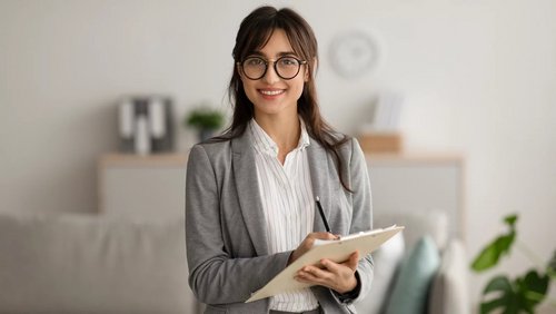 Eine Frau mit Brille in einem grauen Blazer hält ein Notizbuch und lächelt. Im Hintergrund sind Möbel und Pflanzen sichtbar.