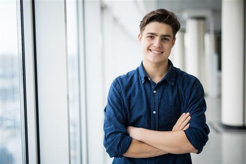 A young man with brown hair stands smiling with his arms crossed in front of a window in a modern building.