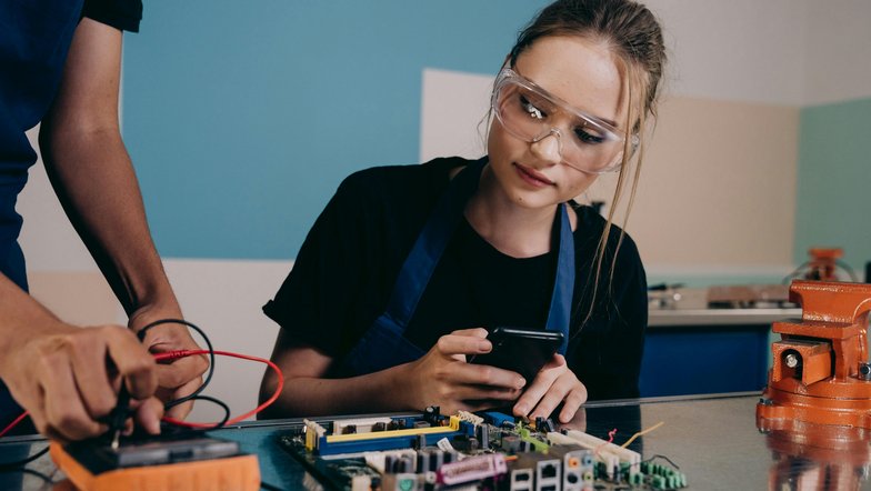 A young woman wearing safety goggles is looking at a smartphone while another person is working on a circuit board. Both are in a technical environment.
