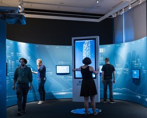 Eine interaktive Ausstellung mit mehreren Personen, die an Bildschirmen stehen. Die Wände sind mit grafischen Darstellungen und Informationen zum Thema Coronavirus dekoriert. Ein zentraler Bildschirm zeigt Daten und Statistiken zur Pandemie.