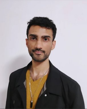 A young man with short, dark hair is wearing a black top and a gold chain. He is looking directly at the camera.