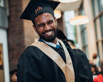 A man in a graduation gown and holding a diploma smiles as he stands in a festive setting.