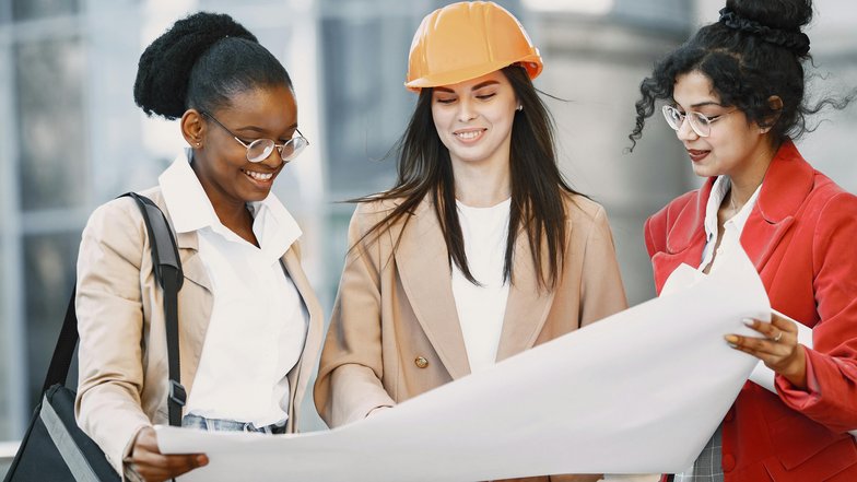 Kategorie Ingeniurwissenschaften SRH University Three women are looking at construction plans together while standing in a modern urban environment. One woman is wearing an orange helmet, while the others are wearing glasses and smiling.