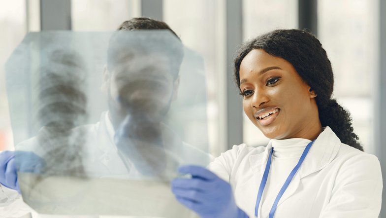 Eine Ärztin betrachtet ein Röntgenbild mit einem Lächeln. Sie trägt einen weißen Laborkittel und blaue Handschuhe, während sie in einem hellen Raum steht.