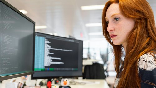 A woman with red hair is intently looking at two computer screens displaying programming code.