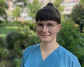 Eine Person mit kurzen, braunen Haaren und einer blauen Arbeitskleidung steht im Freien, umgeben von Pflanzen und Bäumen.