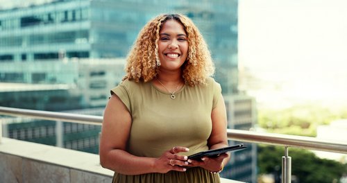 A woman with curly hair is holding a tablet and smiling while standing on a terrace overlooking a city.