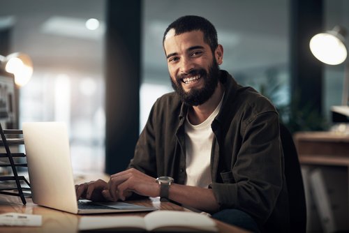A man with a beard is sitting at a desk, smiling as he works on a laptop. A bright office environment is in the background.