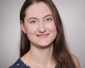Eine Frau mit langen, braunen Haaren und einem blauen Oberteil mit Spitzenmuster lächelt in die Kamera vor einem neutralen Hintergrund.