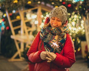 Eine Frau in einem roten Mantel und grauer Mütze hält ein Wunderkerze, umgeben von festlicher Beleuchtung und Dekoration.