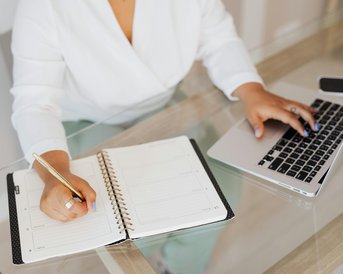A person is writing in a notebook while working on a laptop. The table is made of glass, and the surroundings are bright and modern.