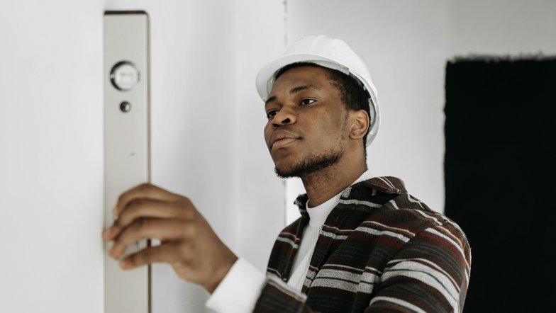 Ein Mann mit einem weißen Helm überprüft mit einer Wasserwaage die Ebenheit einer Wand. Er trägt ein gestreiftes Hemd und konzentriert sich auf seine Aufgabe.