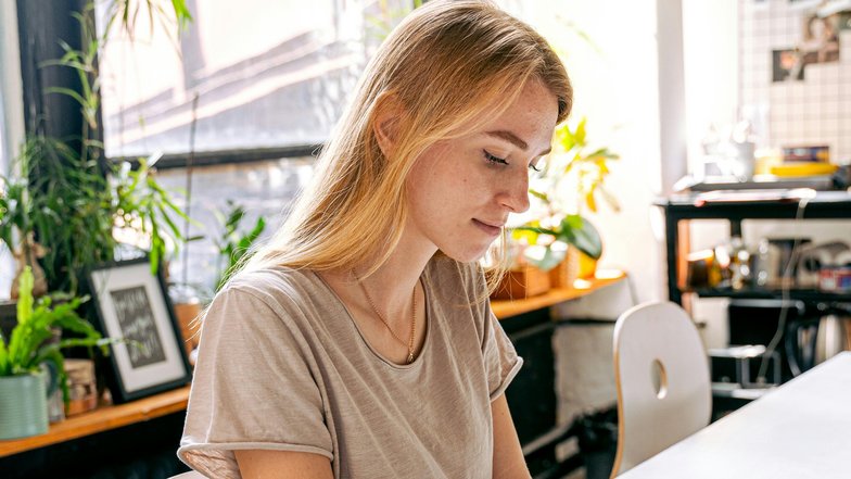 A young woman is sitting at a table in a creative space, drawing with markers on paper. Surrounding her are plants and various art supplies.
