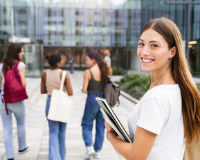 Eine junge Frau mit langen Haaren hält Bücher in der Hand und lächelt, während sie einer Gruppe von drei anderen Frauen in einem modernen Umfeld folgt.