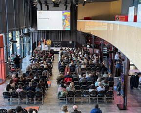 Eine große Menschenmenge sitzt in einem modernen Veranstaltungsraum, während ein Redner auf einer Bühne eine Präsentation hält.