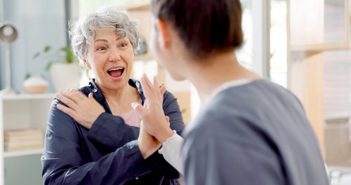 Eine ältere Frau mit grauen Haaren zeigt Freude und Überraschung im Gespräch mit einer jüngeren Person in einem hellen Raum.