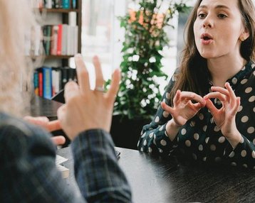 Zwei Frauen sitzen an einem Tisch und unterhalten sich. Eine Frau mit langen, braunen Haaren spricht lebhaft, während die andere Frau aufmerksam zuhört und mit den Händen gestikuliert. Im Hintergrund sind Bücherregale zu sehen.