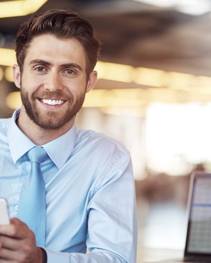 A smiling man in a light blue shirt is holding a smartphone while sitting at a desk with a laptop.