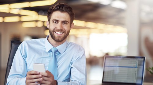 A smiling man in a light blue shirt is holding a smartphone while sitting at a desk with a laptop.