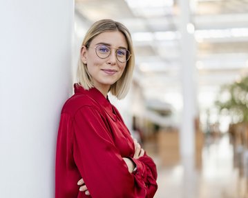 Eine junge Frau mit blonden, schulterlangen Haaren und Brille steht an einer Säule in einem modernen Innenraum. Sie trägt ein rotes Hemd und hat die Arme verschränkt. Der Hintergrund zeigt eine helle, offene Umgebung mit Pflanzen.