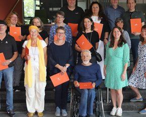 Eine Gruppe von 17 Personen steht auf einer Treppe, einige halten orangefarbene Dokumente. Die Teilnehmer sind unterschiedlich gekleidet und lächeln.
