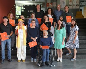 Eine Gruppe von 17 Personen steht auf einer Treppe, einige halten orangefarbene Dokumente. Die Teilnehmer sind unterschiedlich gekleidet und lächeln.