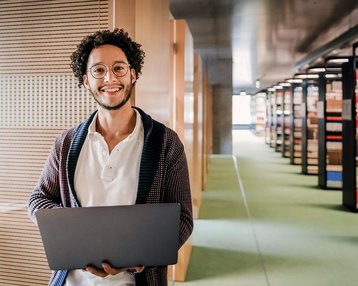 Student mit Brille steht mit seinem Laptop in der Bibliothek