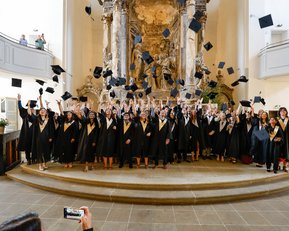Eine Gruppe von Absolventen in schwarzen Roben steht in einer Kirche und wirft ihre Hüte in die Luft, während sie jubeln und feiern. Im Hintergrund ist eine kunstvolle Wandgestaltung zu sehen.