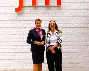 Zwei Frauen stehen vor einer weißen Wand mit dem roten Schriftzug "srh". Die Frau links trägt einen dunkelblauen Anzug mit einem bunten Schal, während die Frau rechts eine schwarz-weiße Jacke und schwarze Hose trägt.