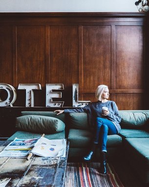 A woman with blonde hair is sitting on a green sofa in a hotel lobby. She holds a cup in her hand and gazes thoughtfully out the window. In the background, the word "HOTEL" is displayed in large letters.