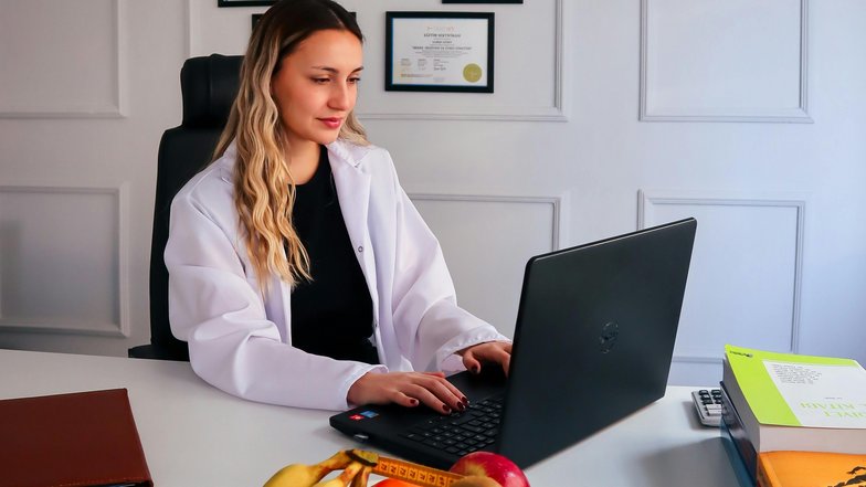 Eine Frau in einem weißen Kittel sitzt an einem Schreibtisch und arbeitet an einem Laptop. Auf dem Tisch befindet sich eine Schüssel mit Obst und ein Maßband. Im Hintergrund hängen Zertifikate an der Wand.